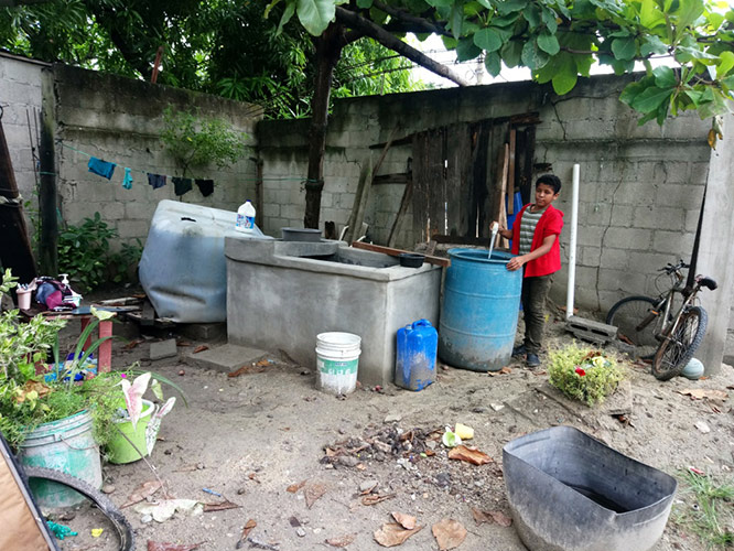 Water storage containers (from left to right): plastic tank with bleach (cisterna), cement tank (pila), bucket (in front of the pila), plastic drum (baril). Clothes are washed on cement washboards like the one at the left side of the pila. The boy is a Monte Verde volunteer checking for mosquito larvae in the plastic drum.